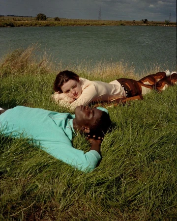 Two people, one Black and one white, lie side-by-side in a grassy field near a lake under a cloudy sky, creating a peaceful outdoor scene.