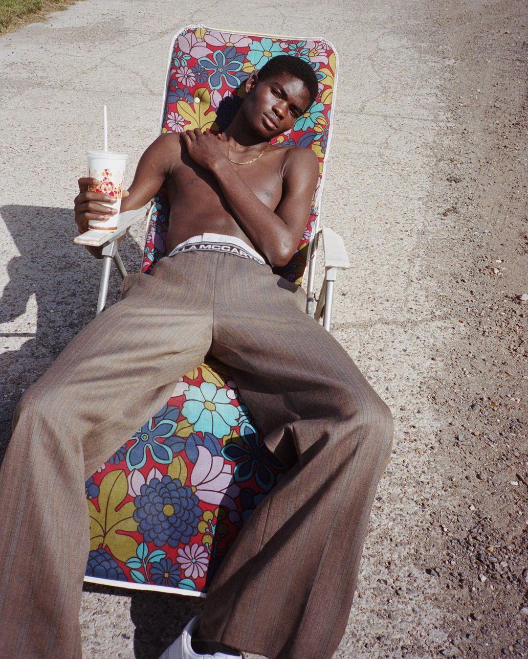 A young Black man sits on a floral beach chair outdoors, wearing brown pants, holding a drink, and relaxing in the sunlight.
