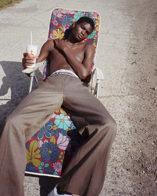 A young Black man sits on a floral beach chair outdoors, wearing brown pants, holding a drink, and relaxing in the sunlight.