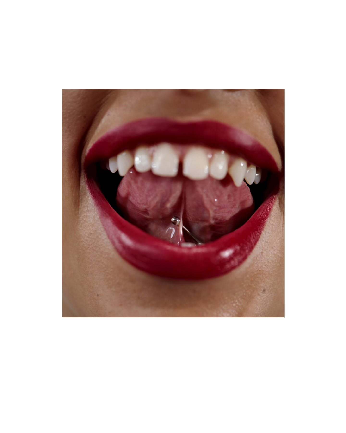 Close-up of an open mouth with red lipstick, white teeth, tongue piercing, and two sections of the tongue.