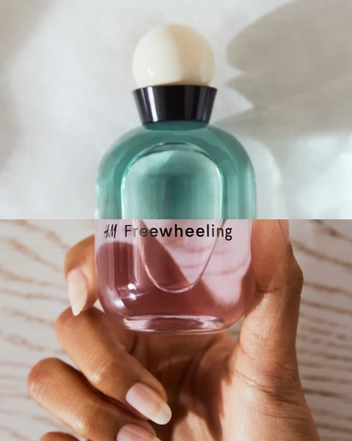 Close-up of a perfume bottle with a white cap, held by a hand with manicured nails against a light background, featuring the H&M Freewheeling logo.