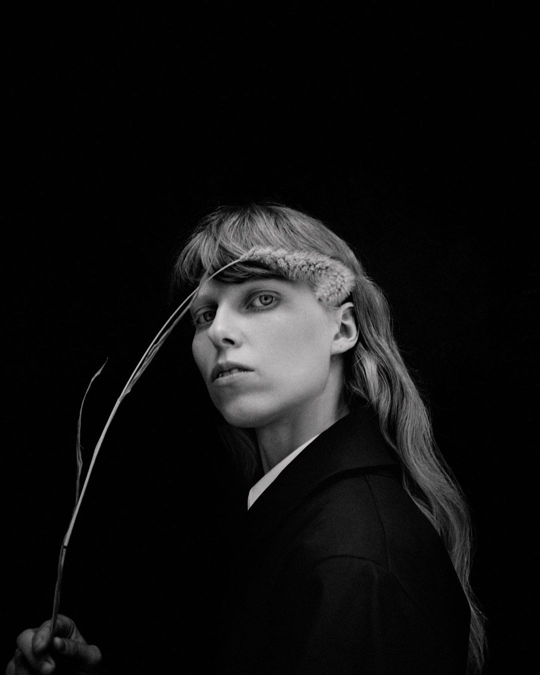 Monochrome portrait of a person with light skin and long hair holding a sprig, set against a black backdrop, exhibiting an editorial style.