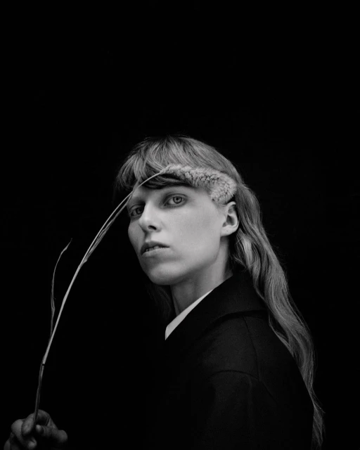 Monochrome portrait of a person with light skin and long hair holding a sprig, set against a black backdrop, exhibiting an editorial style.
