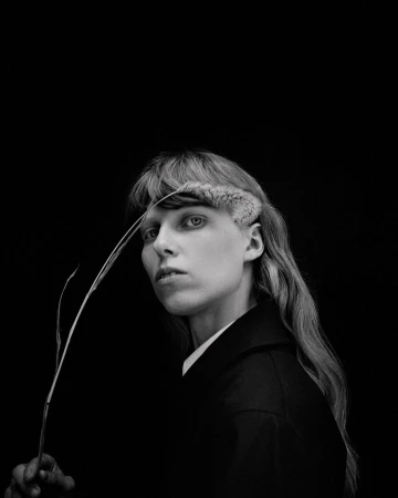 Monochrome portrait of a person with light skin and long hair holding a sprig, set against a black backdrop, exhibiting an editorial style.