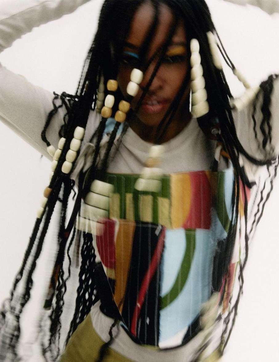 Eye-level shot of a Black woman with beaded braids, wearing a colorful artistic top, against a white backdrop in a studio setting.