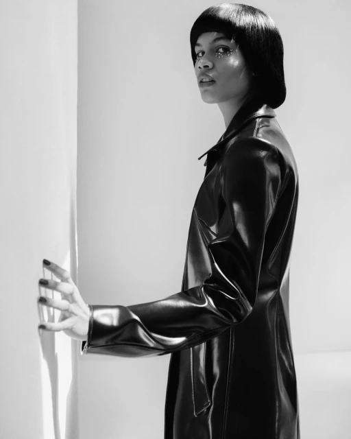 Striking black and white portrait of a woman in a glossy black trench coat with glittering makeup posing against a column.