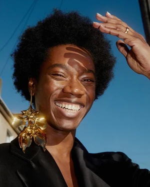 Smiling Black man wearing a large gold statement earring and a black blazer against a clear blue sky by Johanna Nyholm