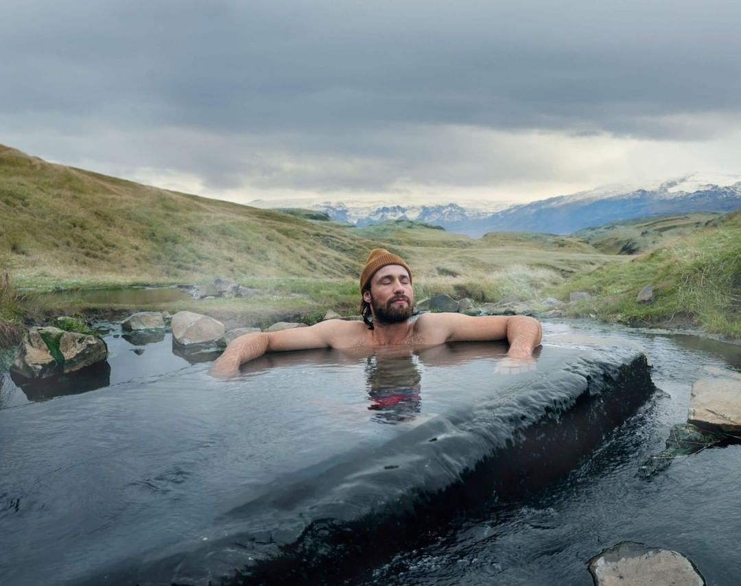 Man soaks in geothermal hot spring in Iceland with snow-capped mountains and cloudy sky in the background; relaxation and travel concept.