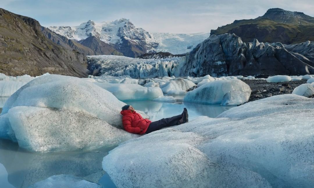 Adult male in red jacket relaxing on glacier ice in Jokulsarlon, Iceland, with mountains and glacial lagoon in background under daylight sky.