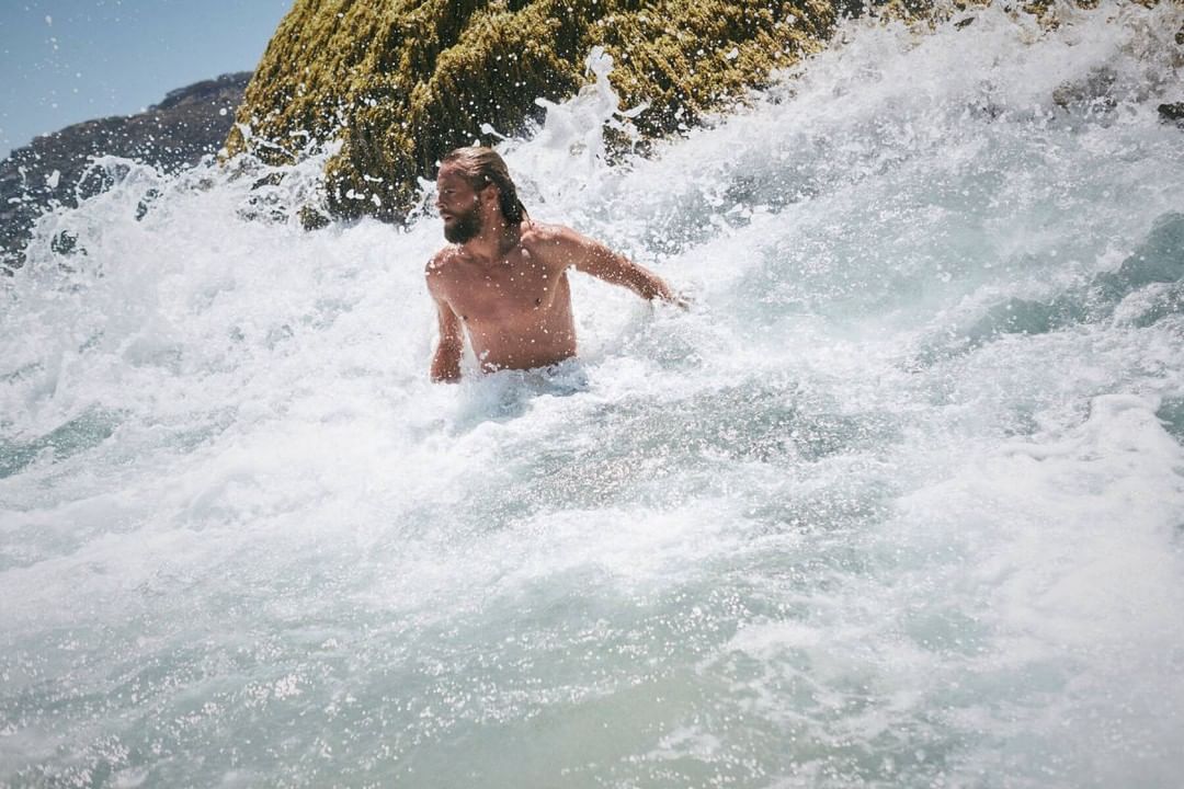 An adult male with long hair stands waist-deep in white foamy ocean waves with a rocky cliff covered in green algae in the background.