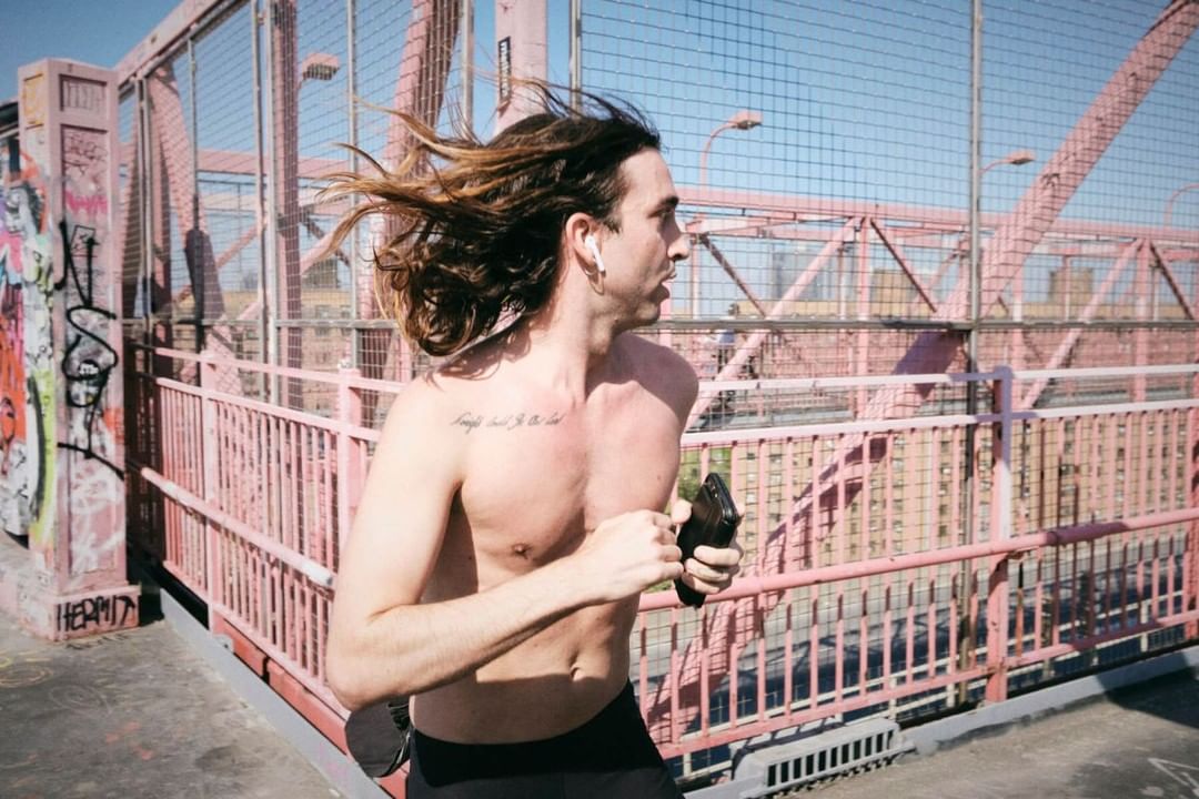 Athletic adult male running shirtless across an urban bridge, holding a phone, with city structures in the background under bright sunlight.