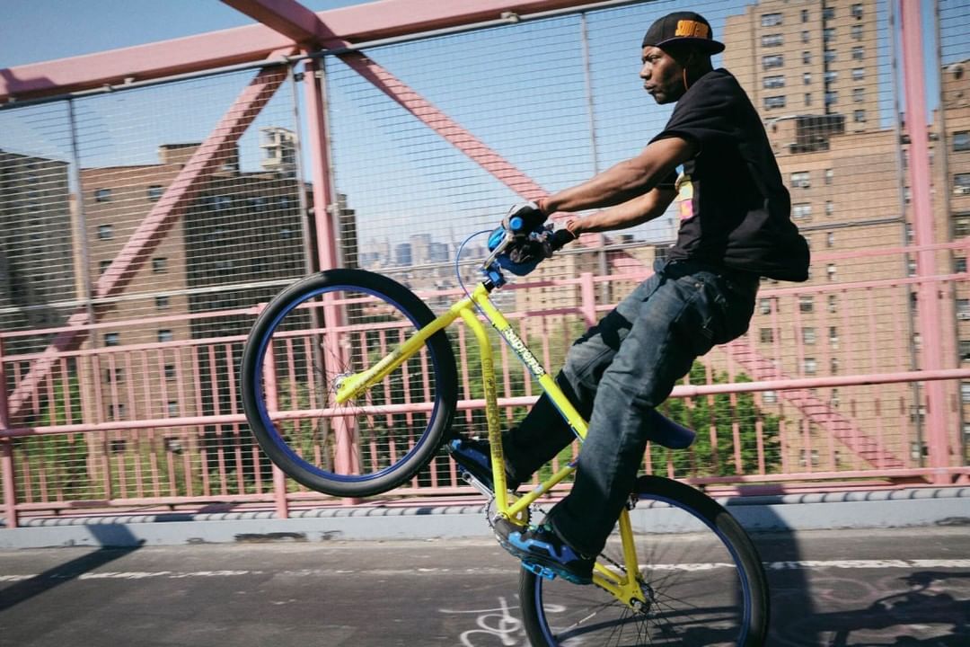 Adult male performs a wheelie on a yellow and blue bicycle on a pink bridge, with a city building backdrop, during daylight hours.