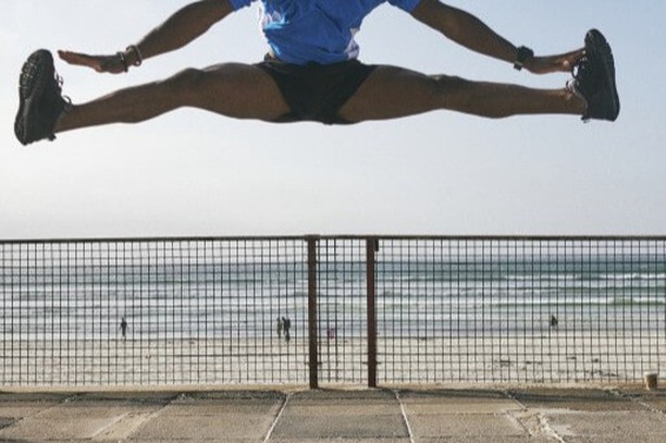An athletic man is leaping high above a metal fence, showcasing a wide split in the air against a beach and ocean backdrop.