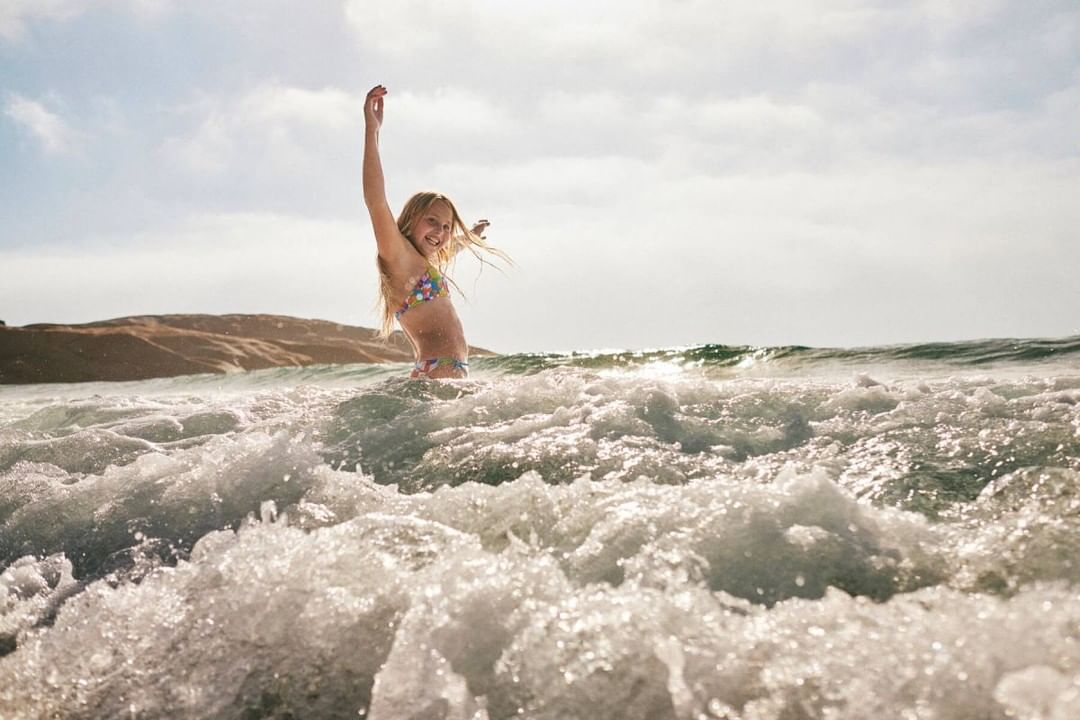 Young girl with blonde hair in a colorful swimsuit standing in ocean water with her arms raised, smiling on a sunny day at the beach.