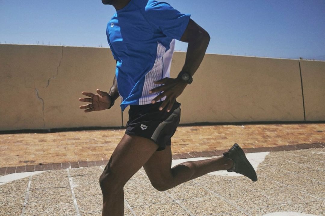 Side view of a dark-skinned adult male running outdoors in a blue and white shirt, black shorts, and black shoes against a clear sky.