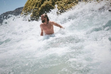 Man with beard swimming in crashing ocean waves near a rocky shore with sea foam spray