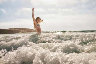 A young girl in a patterned bikini stands waist-deep in churning ocean waves, raising one arm joyfully.