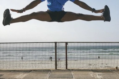 Athlete caught mid air jumping with split legs against a metal mesh fence and ocean beach background.