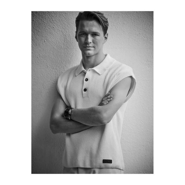 Black and white portrait of a young man in a sleeveless knitted shirt, with arms crossed and a watch on his left wrist, against a textured wall.