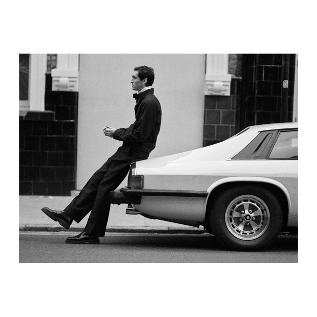 Monochrome image of a male model in dark attire seated on a vintage white car, captured in an urban setting with diffused natural light.