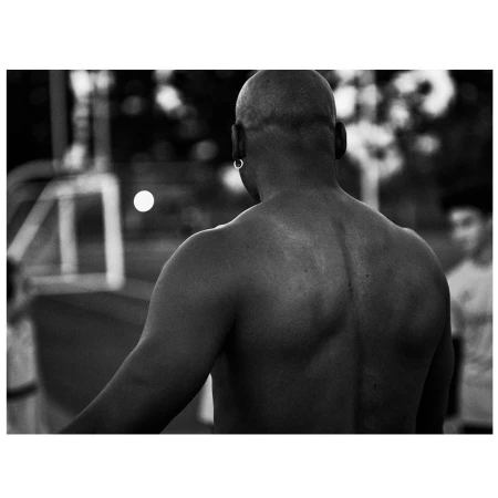 Black and white portrait of a bald man, seen from the back with his bare upper body and hoop earring, in an outdoor setting.