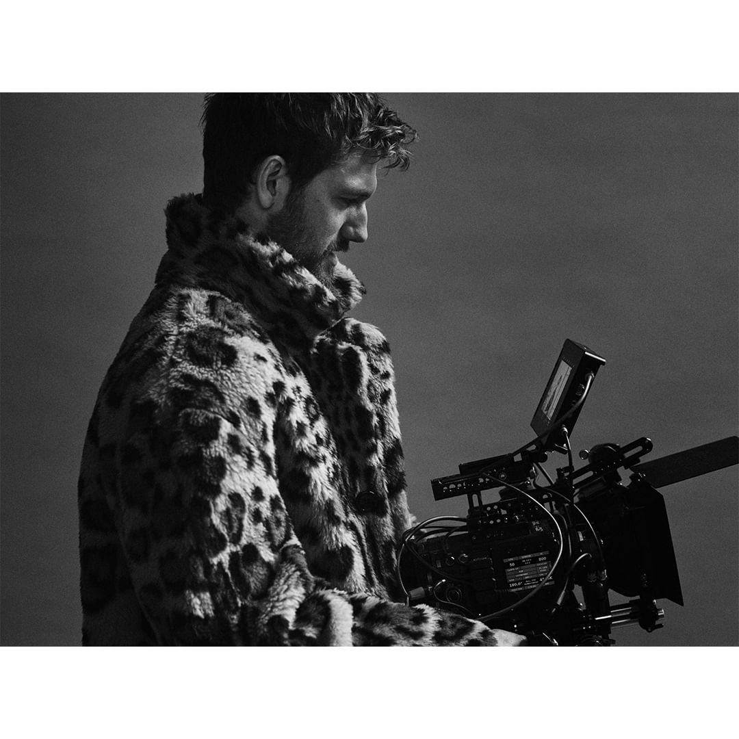 Man in leopard print coat operates professional film camera against grey backdrop in a black and white studio setting.