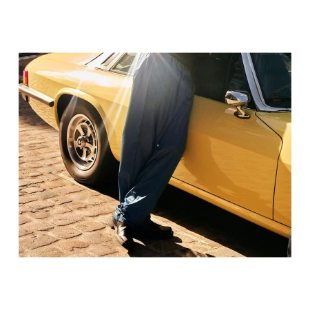 Partial view of a yellow vintage car on a brick surface with a person wearing blue pants leaning against the car.