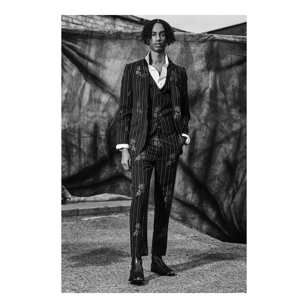 Monochrome portrait of a Black male model in a floral pinstripe suit with an open white collared shirt, standing outdoors.