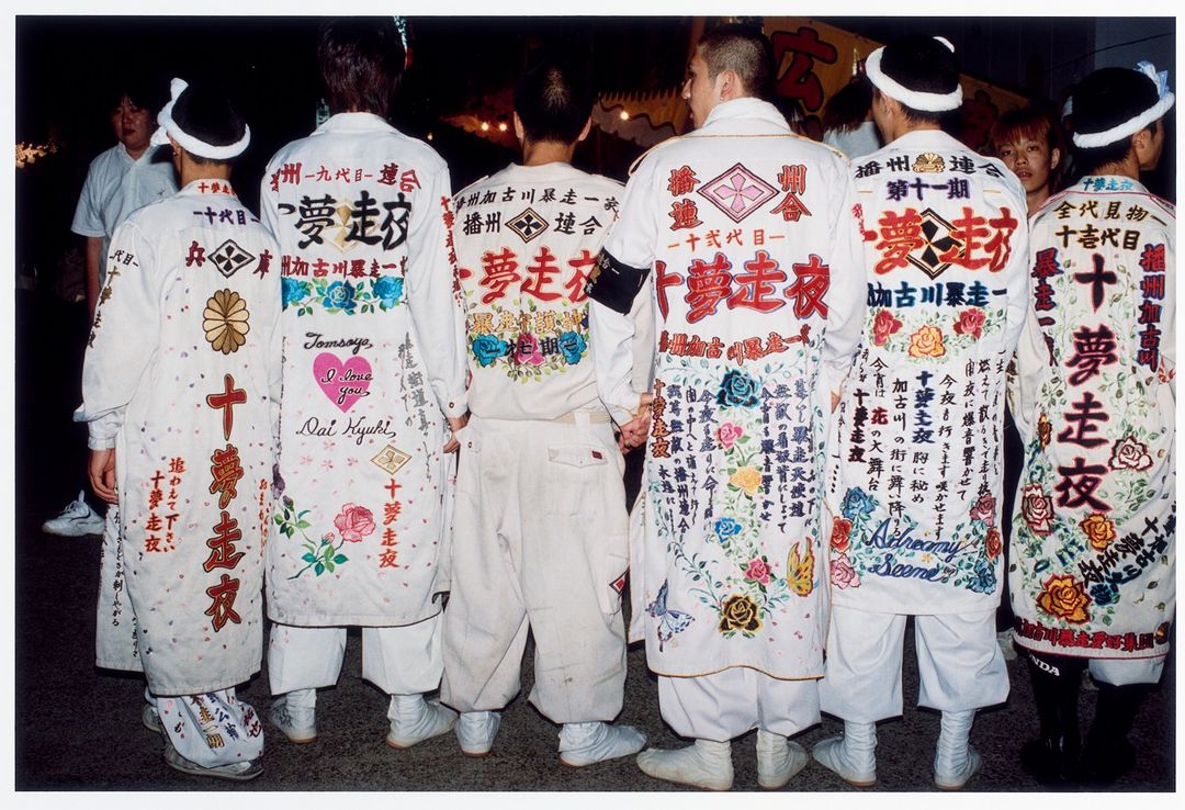 A group of six people with white coats covered in text and graphics stand together outdoors, showcasing a blend of fashion and culture.