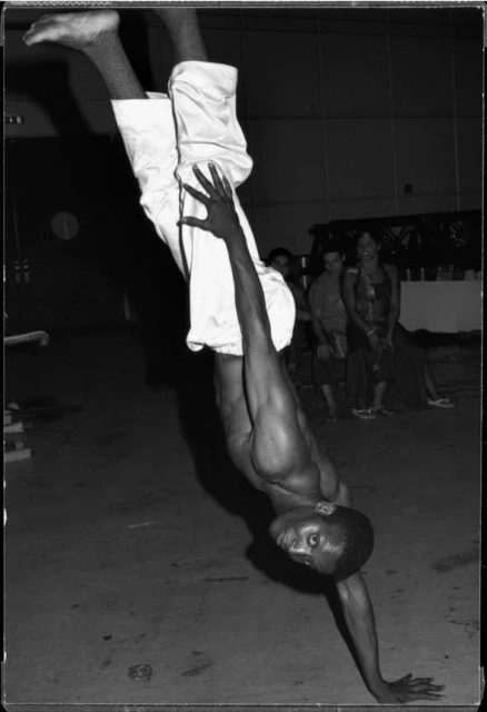 An athletic adult male in white pants doing a handstand indoors, with a group of people in the background. Black and white photography.