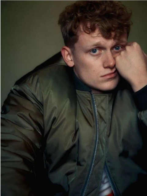 Portrait of an adult male with red curly hair, wearing a green bomber jacket against an olive green background in soft studio lighting.