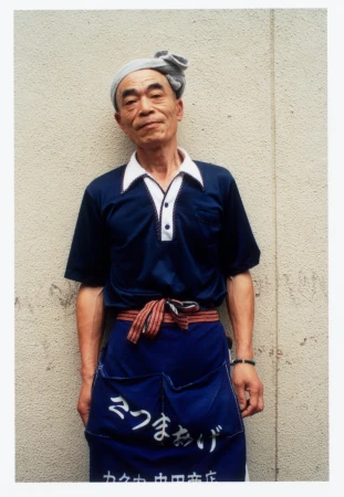 Older Asian man in uniform and headwrap standing in front of textured wall, with apron. Japanese shopkeeper portrait.