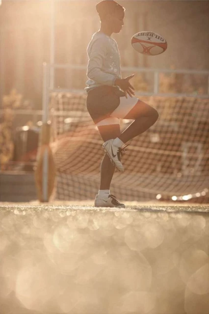 A young adult male in sportswear skillfully kicks a rugby ball in a sunny outdoor setting with a net and a football goalpost in the background.