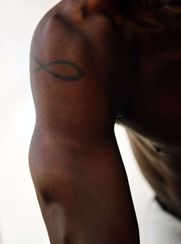 Close-up on the arm and shoulder of an adult with dark skin, showing muscle definition and a black tattoo against a plain white background.