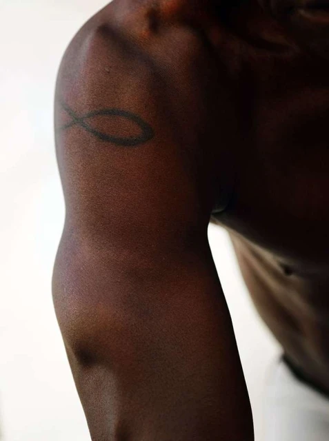 Close-up on the arm and shoulder of an adult with dark skin, showing muscle definition and a black tattoo against a plain white background.