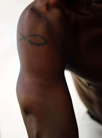 Close-up on the arm and shoulder of an adult with dark skin, showing muscle definition and a black tattoo against a plain white background.