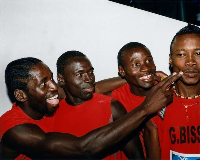 Four adult male athletes wearing red tank tops pose indoors, smiling and pointing. Head and shoulder shot with a plain white background.