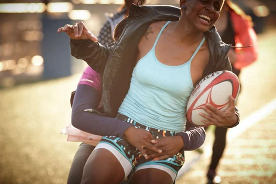 Two women smiling and embracing on a sports field, one holding a rugby ball, captured in a medium shot with natural lighting.