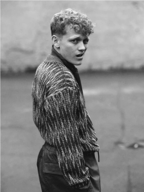 Monochrome portrait of a young adult male with curly hair, wearing a patterned sweater, looking over his shoulder outdoors.