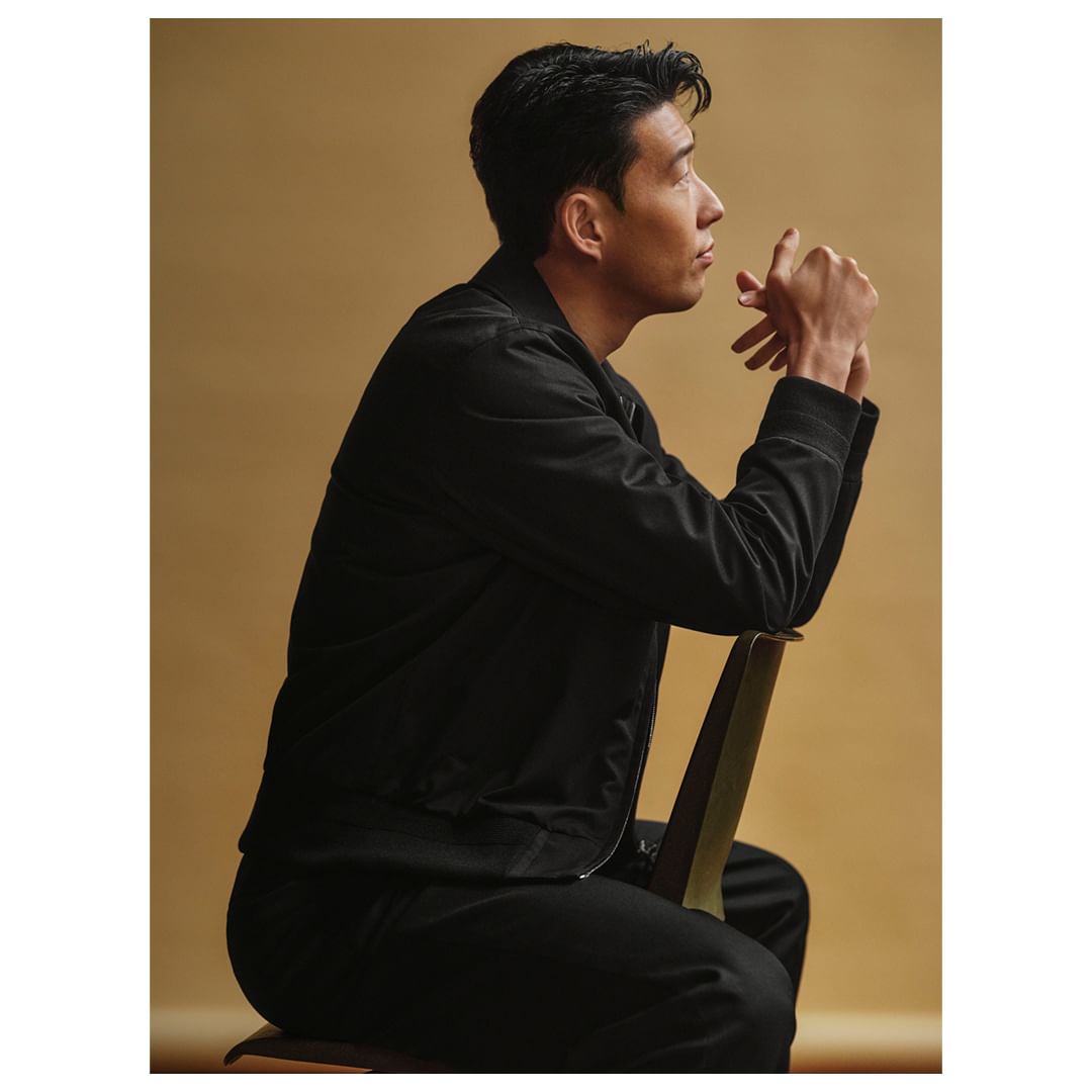 Portrait of an adult Asian male seated on a chair, gazing away, wearing a black jacket against a tan backdrop in a photo studio.