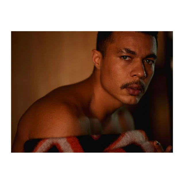 Portrait of a bare-chested adult male model with a moustache, facing camera, holding a patterned blanket, in an indoor environment.