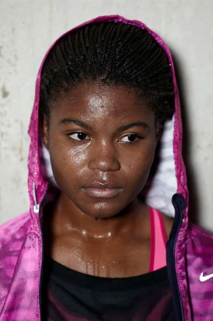 Close-up portrait of a sweaty woman with dark skin and braided hair, wearing a pink hooded jacket in a bright indoor setting.