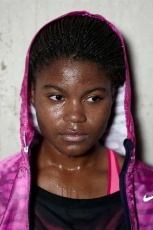 Close-up portrait of a sweaty woman with dark skin and braided hair, wearing a pink hooded jacket in a bright indoor setting.