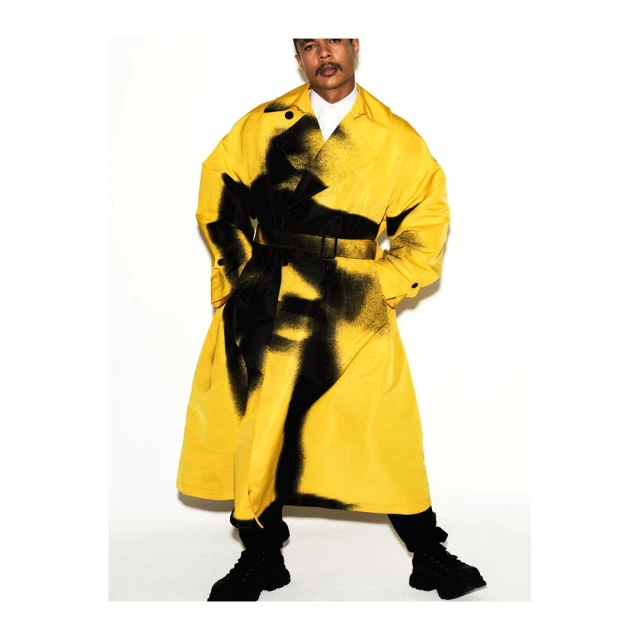 Fashionable man poses in yellow trench coat with black graphic design against a white backdrop in a studio, showcasing style.