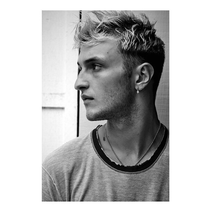 Monochrome portrait of a young adult male with blond-streaked hair looking left; the man is wearing a t-shirt and a ball chain necklace.