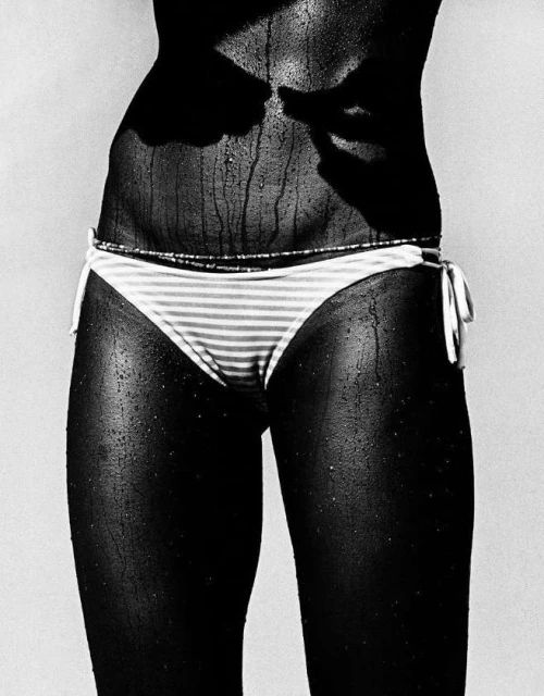 Close-up black and white shot of a woman's midsection with striped bikini bottom, sunlit with visible water droplets on her skin.