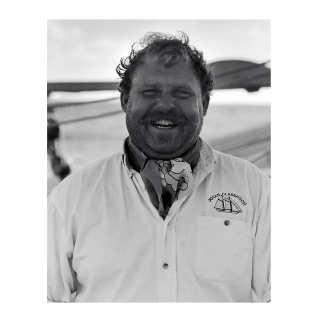 Monochrome portrait of a smiling adult male with curly hair and mustache, wearing a white shirt and patterned neckerchief outdoors.