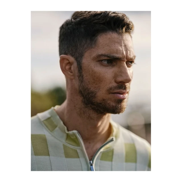 Portrait of a man with short brown hair and a light beard, wearing a light green and white checked shirt, gazing slightly off-camera outdoors.