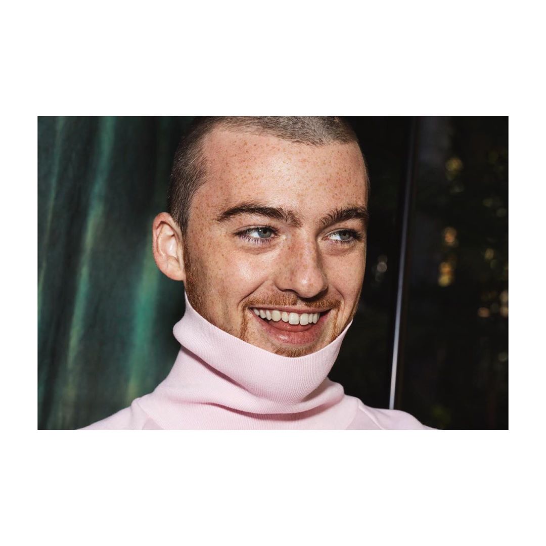 Close-up studio portrait of a young adult male smiling and wearing a pink turtleneck, set against a dark green curtain backdrop.