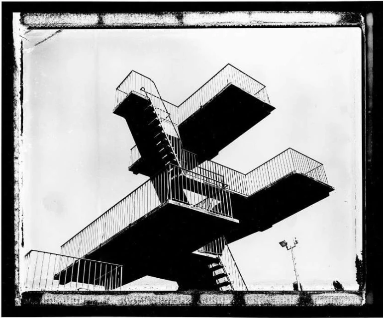Monochrome photograph of a high diving platform with stairs and railings against a bright sky, showcasing its architectural structure.